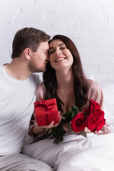Mann küsst glückliche Frau mit Rosen und Blumen im verschwommenen Vordergrund im Schlafzimmer — Stockfoto