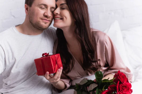 Smiling woman holding present and roses near husband in bedroom — Stock Photo