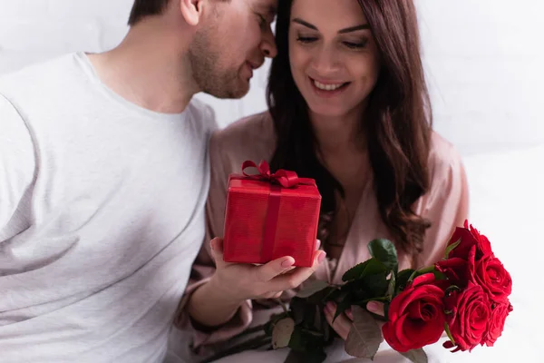 Geschenk und Blumen in den Händen einer glücklichen Frau in der Nähe ihres Mannes zu Hause — Stockfoto