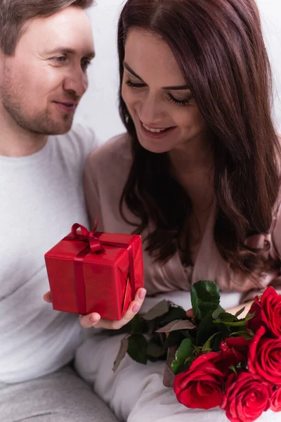 Smiling woman holding gift box and roses near husband in bedroom — Stock Photo