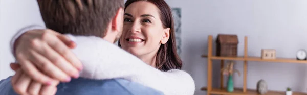 Femme souriante étreignant mari au premier plan flou à la maison, bannière — Photo de stock