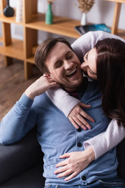 Morena mujer abrazando alegre marido en sofá en casa - foto de stock