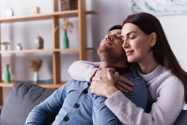 Brunette woman with closed eyes hugging husband sitting on couch — Stock Photo