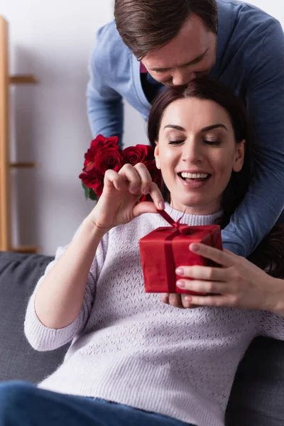 Mujer sonriente sosteniendo arco de caja de regalo cerca de marido con rosas en casa - foto de stock
