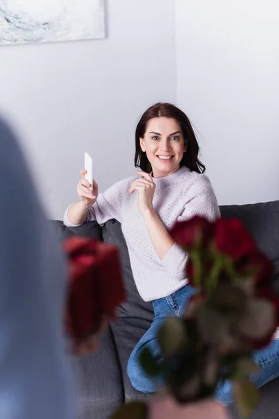 Lächelnde Frau hält Handy neben Ehemann mit Blumen und Geschenk auf verschwommenem Vordergrund — Stockfoto