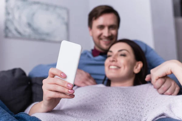 Смартфон в руке улыбающейся женщины, лежащей на диване рядом с мужем — стоковое фото