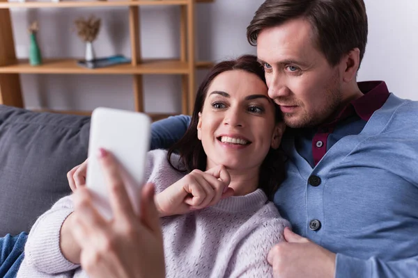 Positive Frau hält Handy im verschwommenen Vordergrund neben Ehemann im Wohnzimmer — Stockfoto