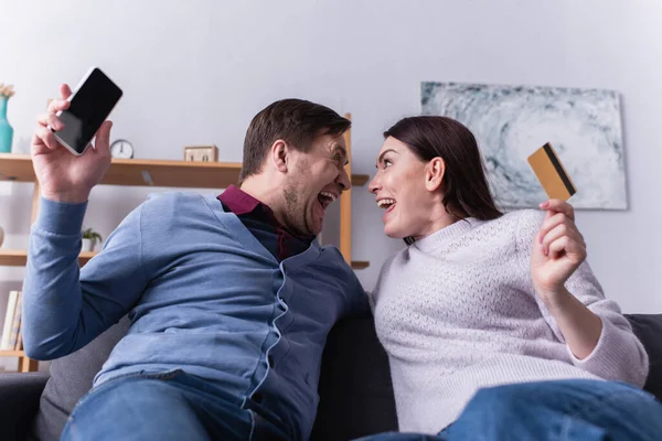 Casal animado olhando uns para os outros enquanto segurando smartphone e cartão de crédito — Fotografia de Stock