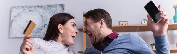 Amazed couple with credit card and smartphone looking at each other at home, banner — Stock Photo