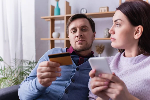 Hombre mirando la tarjeta de crédito cerca de la esposa con teléfono inteligente en la sala de estar - foto de stock