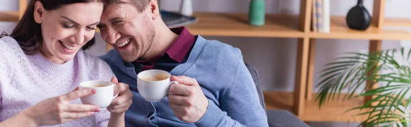 Alegre pareja adulta sosteniendo tazas de café en casa, pancarta - foto de stock