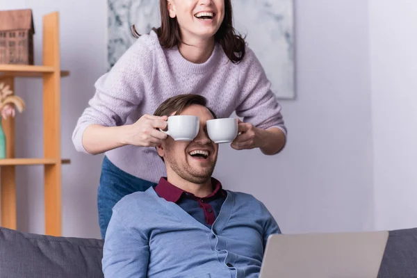Cheerful woman covering face of husband with cups of coffee at home — Stock Photo