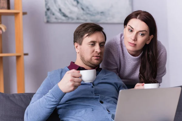 Uomo con tazza guardando laptop vicino moglie — Foto stock