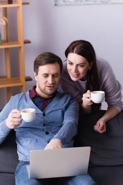 Mujer sonriente con taza mirando portátil cerca de marido en sofá - foto de stock