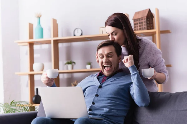 Aufgeregter Mann mit Tasse blickt auf Laptop neben Ehefrau — Stockfoto