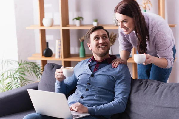 Homem usando laptop e olhando para a esposa com copo na sala de estar — Fotografia de Stock