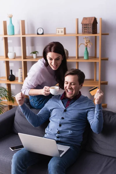 Возбужденный мужчина держит кредитную карту возле ноутбука и жена с чашкой дома — стоковое фото