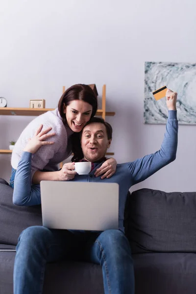 Mujer con taza abrazando marido con tarjeta de crédito cerca de la computadora portátil en primer plano borrosa - foto de stock