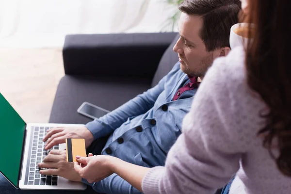 Hombre usando el ordenador portátil con pantalla verde cerca de la esposa con tarjeta de crédito y taza en primer plano borrosa - foto de stock