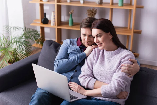 Sorrindo mulher olhando para laptop perto do marido no sofá — Fotografia de Stock