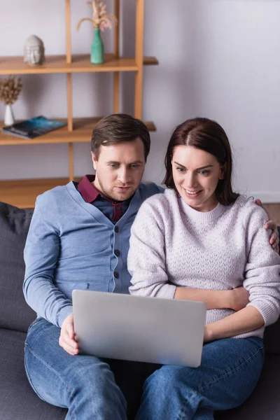 Hombre mirando el ordenador portátil cerca de esposa alegre en el sofá en la sala de estar - foto de stock