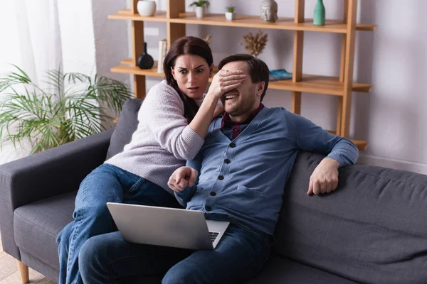 Mulher assustada cobrindo os olhos do marido apontando para o laptop no sofá — Fotografia de Stock