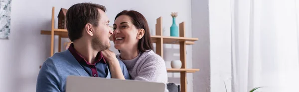 Mulher sorrindo olhando para o marido perto do laptop em primeiro plano borrado em casa, banner — Fotografia de Stock