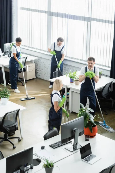 Hohe Blickwinkel auf multiethnische Reinigungskräfte Waschboden in der Nähe von Computern im Büro — Stockfoto