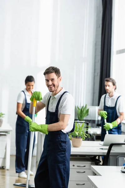 Lächelnde Putzfrau in Gummihandschuhen hält Wischmopp neben Kollegen auf verschwommenem Hintergrund im Büro — Stockfoto