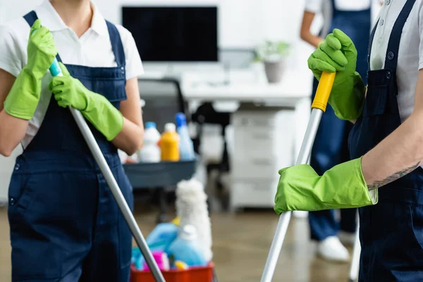 Cropped view of cleaners in rubber globes holding mops in office — Stock Photo