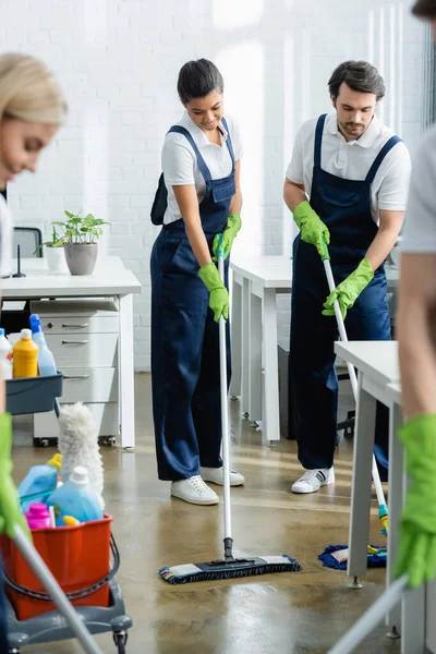 Limpiadores multiétnicos lavando el piso con trapeadores cerca de colegas en primer plano borroso en la oficina - foto de stock