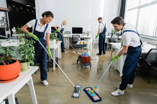 African american cleaner smiling while washing floor near colleague in office — Stock Photo