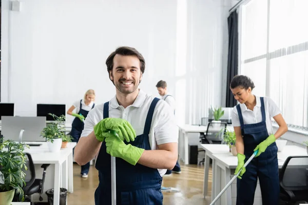 Limpiador con fregona sonriendo a cámara cerca de colega afroamericano en la oficina sobre fondo borroso - foto de stock