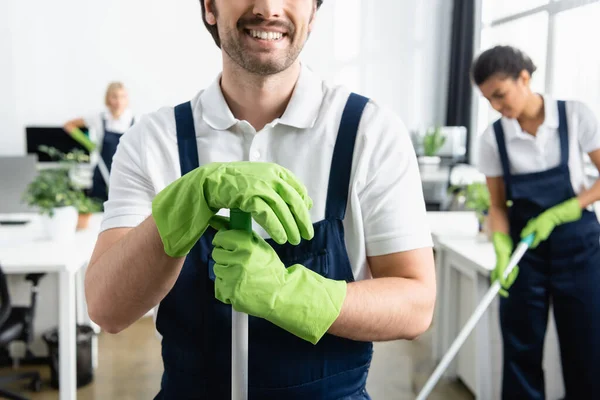 Trabajador sonriente de la empresa de limpieza sosteniendo fregona y sonriendo en la oficina - foto de stock