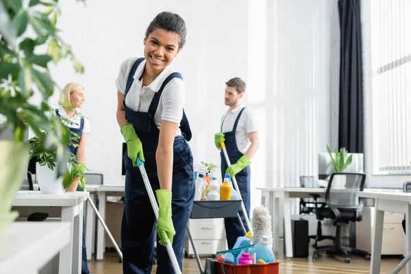 Afroamerikanische Putzfrau hält Wischmopp in der Nähe von Kollegen und Fabrik — Stock Photo