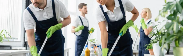 Limpiadores interraciales sosteniendo trapeadores cerca de colegas en un fondo borroso en la oficina, pancarta - foto de stock