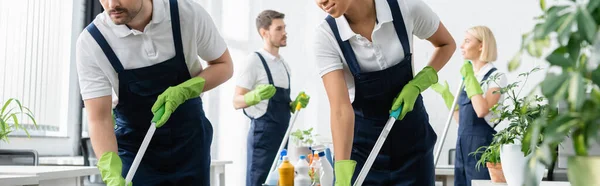 Limpadores inter-raciais segurando esfregonas perto de colegas em fundo turvo no escritório, banner — Fotografia de Stock