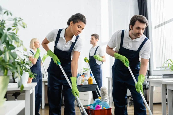 Nettoyants multiethniques tenant des vadrouilles tout en travaillant près de collègues au bureau — Photo de stock