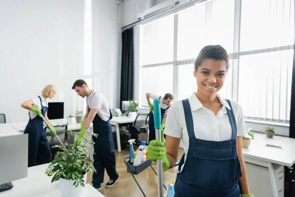 African american cleaner holding mop and smiling at camera while colleagues working in office — Stock Photo