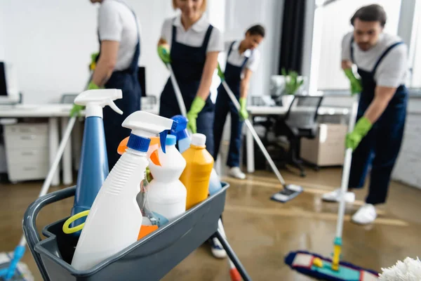 Garrafas com detergentes perto de limpadores trabalhando em fundo desfocado no escritório — Fotografia de Stock