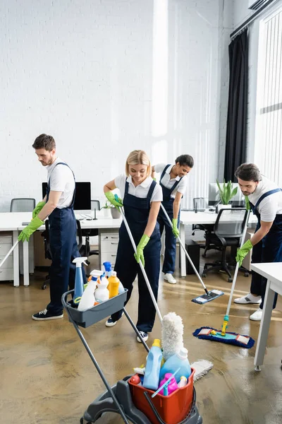 Lächelnde Reinigungskräfte waschen Boden in der Nähe von Waschmitteln im Büro — Stockfoto