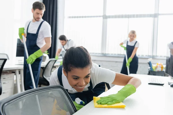 Smiling african american cleaner cleaning table with rag in office — Stock Photo