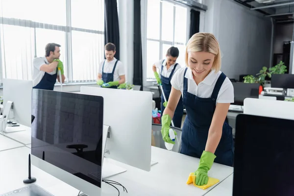 Mesa limpiadora sonriente con detergente cerca de colegas multiétnicos y computadoras en la oficina - foto de stock