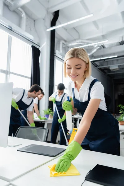 Trabajador positivo de la empresa de limpieza mesa de limpieza con trapo cerca de colegas sobre fondo borroso en la oficina - foto de stock