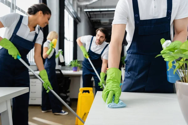 Reiniger Putztisch in Werksnähe und multiethnische Kollegen auf verschwommenem Hintergrund im Büro — Stockfoto