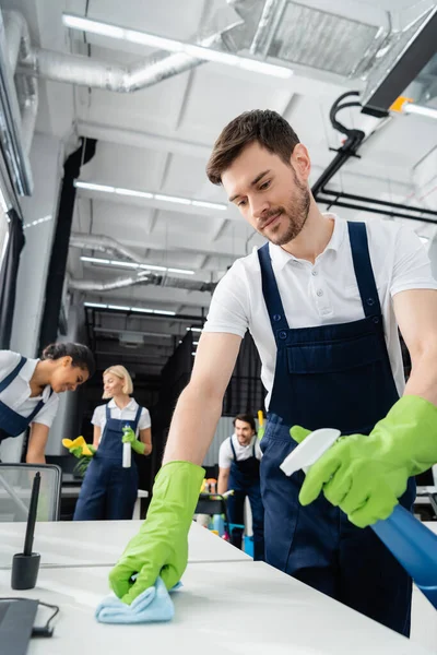 Trabajador de servicio de limpieza mesa de limpieza con detergente cerca de colegas multiétnicos que trabajan en la oficina - foto de stock