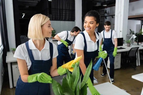 Exotische Reinigungskräfte lächeln einander im Büro an — Stockfoto