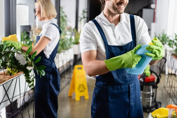 Улыбающийся чистильщик держит моющее средство рядом с коллегой на размытом фоне в офисе — стоковое фото
