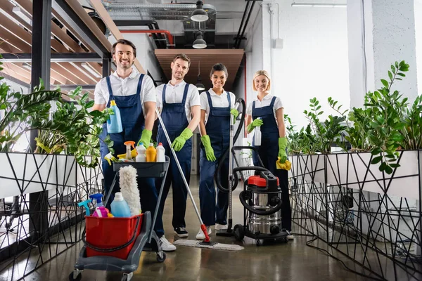 Limpiadores multiétnicos con artículos de limpieza sonriendo a la cámara en la oficina - foto de stock