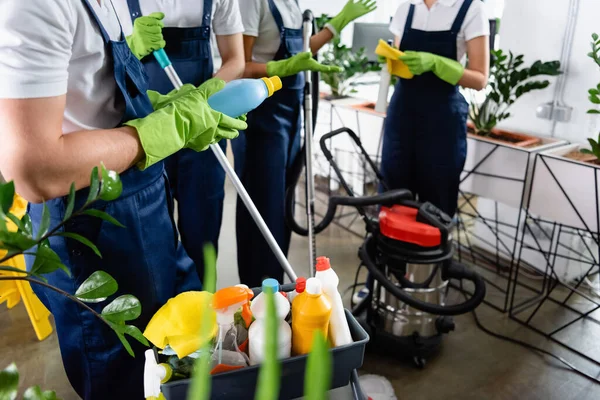 Cropped view of cleaner in rubber gloves holding detergent near colleagues and vacuum cleaner in office — Stock Photo